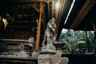 Ilustrasi: Patung penjaga batu berukir di pura Bali dengan latar ukiran kayu keemasan dan pepohonan hijau.