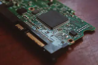 Ilustrasi: Papan sirkuit elektronik hijau dengan mikrocip dan konektor emas di atas permukaan kayu.