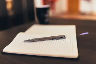 Ilustrasi: Buku catatan bergaris terbuka dengan pulpen perak di atas meja kayu.
