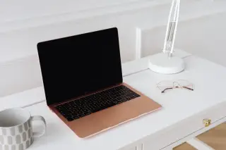 Ilustrasi: Laptop terbuka di atas meja kerja putih berdampingan dengan lampu meja, kacamata, dan cangkir.