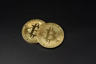 Ilustrasi: Dua keping koin emas Bitcoin bertumpuk di atas permukaan hitam polos.