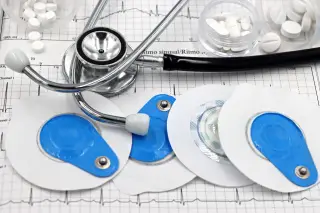 Ilustrasi: Stetoskop, pil obat, dan elektroda EKG tertata di atas kertas grafik jantung.