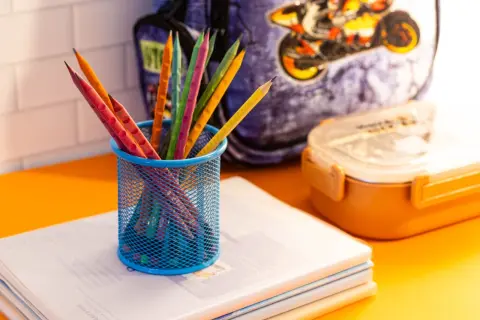 Ilustrasi: Pensil warna di dalam wadah biru di atas buku catatan dengan latar tas dan kotak makan.