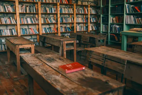 Ilustrasi: Deretan meja dan bangku kayu di dalam ruangan perpustakaan dengan rak buku penuh.