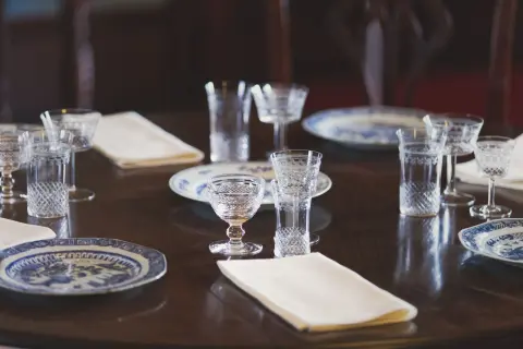 Ilustrasi: Meja makan bundar tertata dengan gelas kristal, serbet, dan piring keramik bermotif biru.