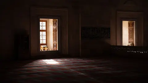 Ilustrasi: Interior masjid sunyi dengan cahaya matahari menyinari karpet bermotif dan dinding batu berkaligrafi.