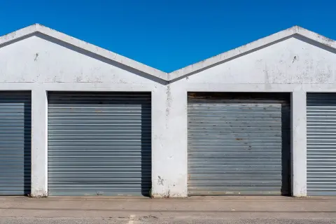 Ilustrasi: Deretan pintu gulung besi pada bangunan gudang putih di bawah langit biru cerah.
