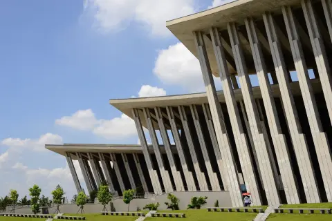 Ilustrasi: Bangunan modern dengan deretan pilar beton besar di bawah langit biru cerah berawan.