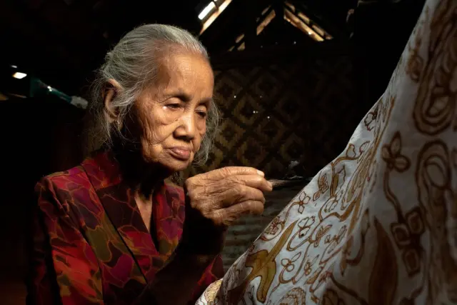 Ilustrasi: Seorang wanita lansia sedang membatik dengan canting pada kain putih bermotif.